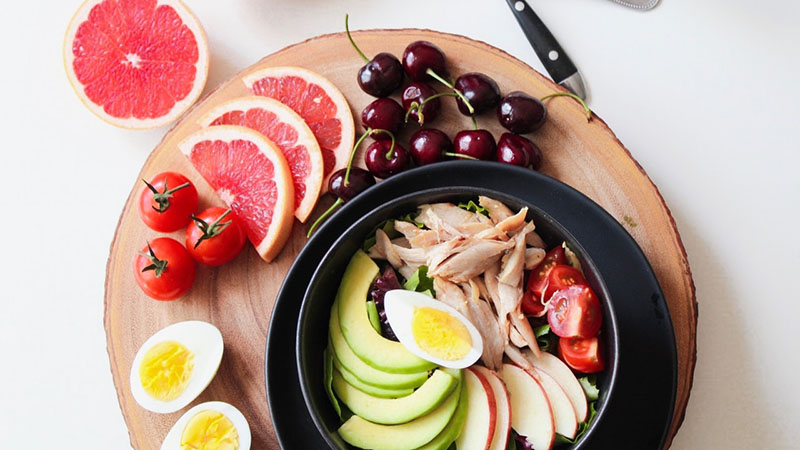 A bowl of chicken salad with cherries, cherry tomatoes, grapefruit and egg on the side.