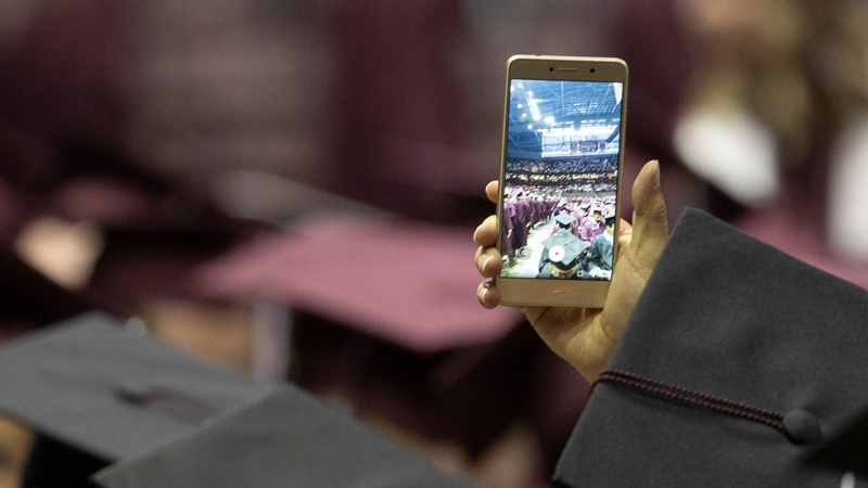 A graduate takes a photo of commencement on his phone.