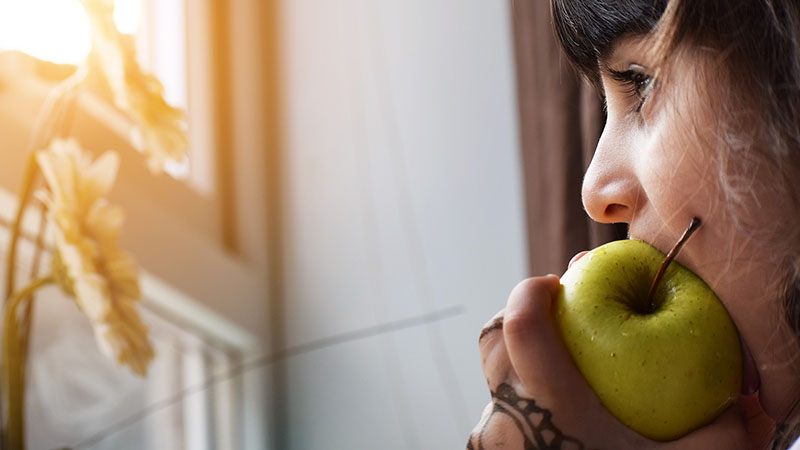 A woman biting on a green apple while looking out the window.