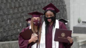 Two graduates wearing masks proudly display their diploma covers after commencement.