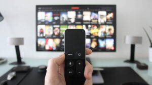 A hand holding a TV remote control and pointing at a TV.