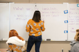Teacher writes on white board in front of classroom.