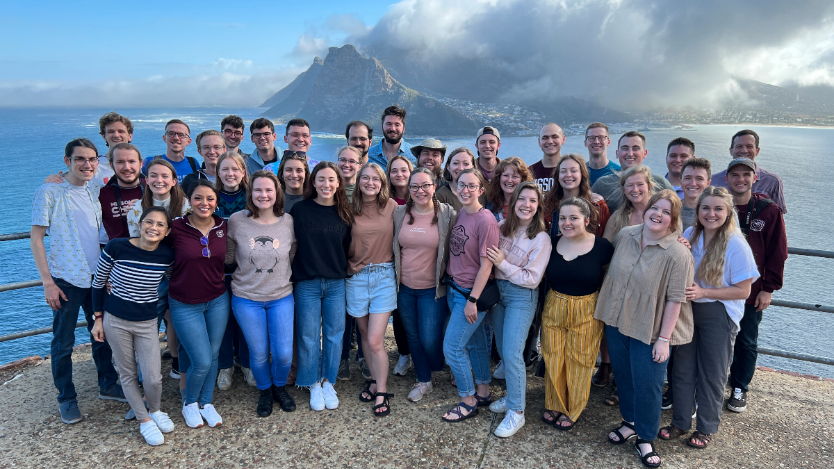 Group poses with sea and mountain in background.