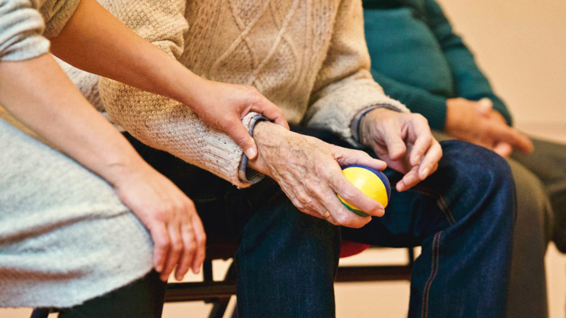 An elderly person holding a stress ball while a younger person offers support.