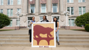 Three women stand in front of Carrington Hall with a Bear Bridge sign.
