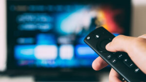 A close-up of a hand holding a remote with a blurred-out TV in the background.