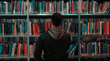 A person wearing a black and grey hoodie looking at a bookshelf.