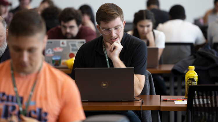 A male student sits thinking at a laptop in a room full of people