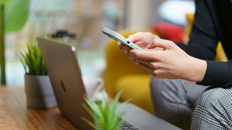 A person using a cell phone while sitting in front of a laptop.