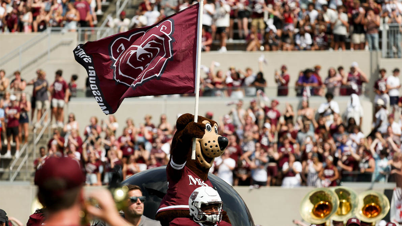 Boomer Bear representing Missouri State University waves a flag at a home football game, with fans and the pride band in the background.