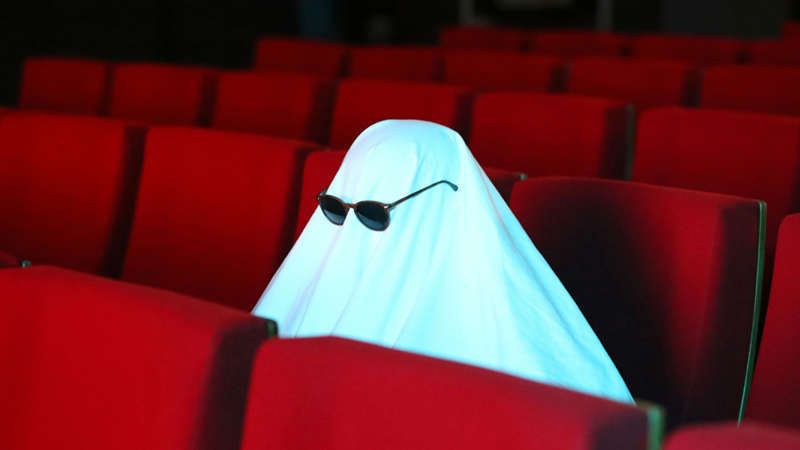 A person in a ghost costume with sunglasses sits in a movie theater with red seats.