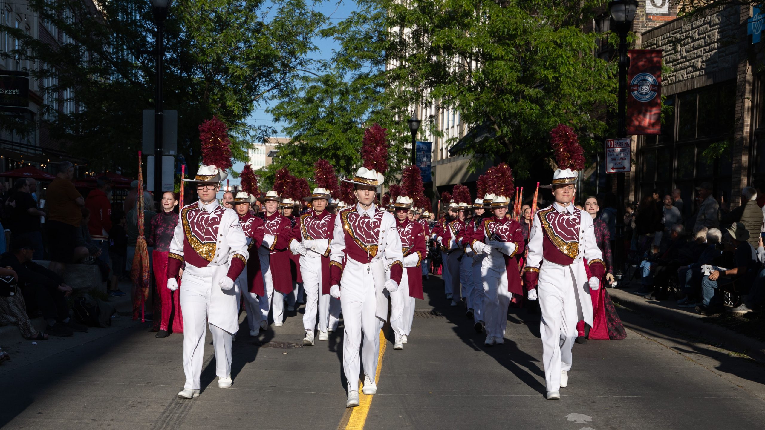 A marching band in white and maroon uniforms parades down a sunlit city street lined with spectators and trees.