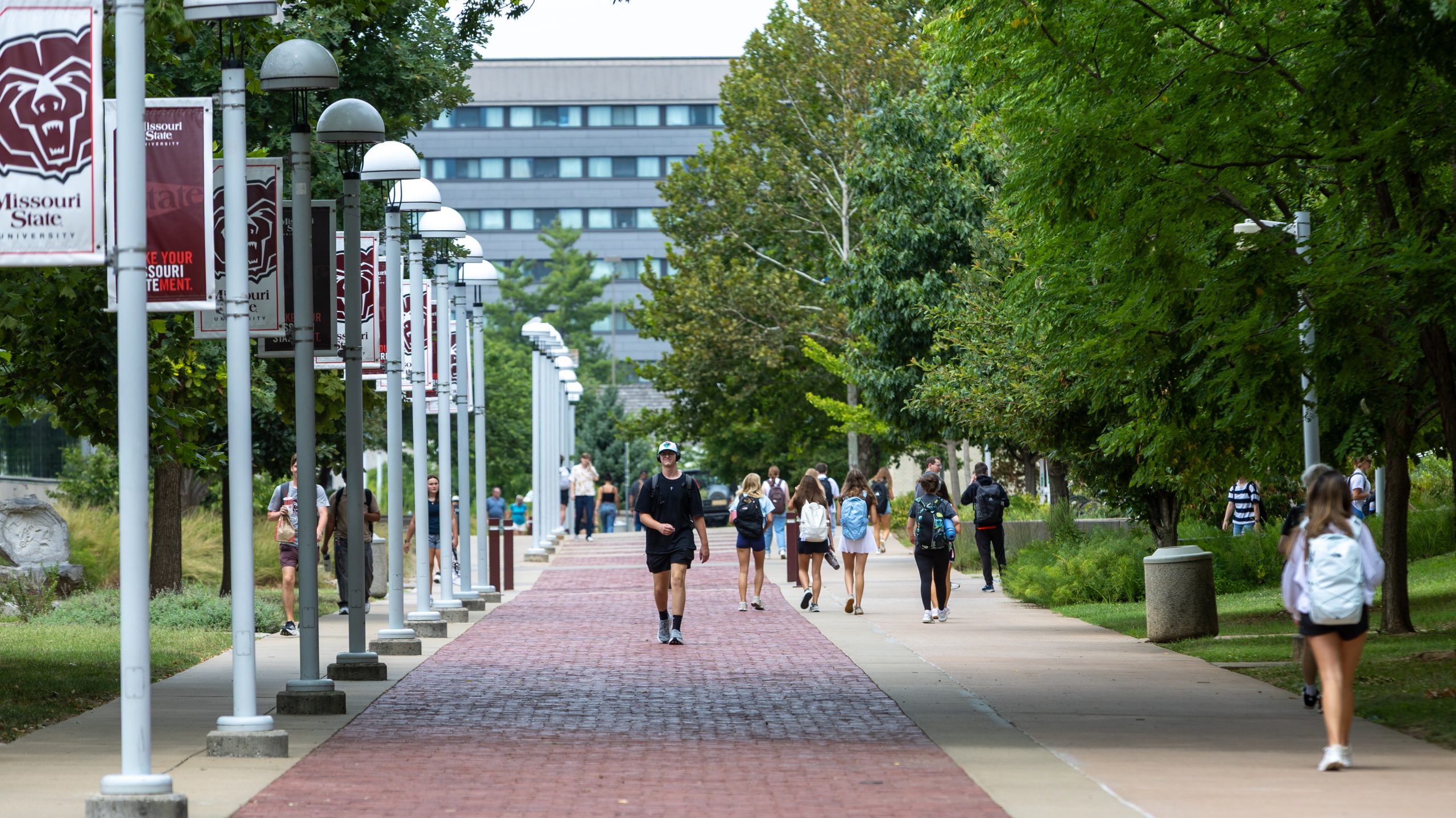 Students stroll along a tree-lined pathway on a university campus, with banners and modern buildings in the background.