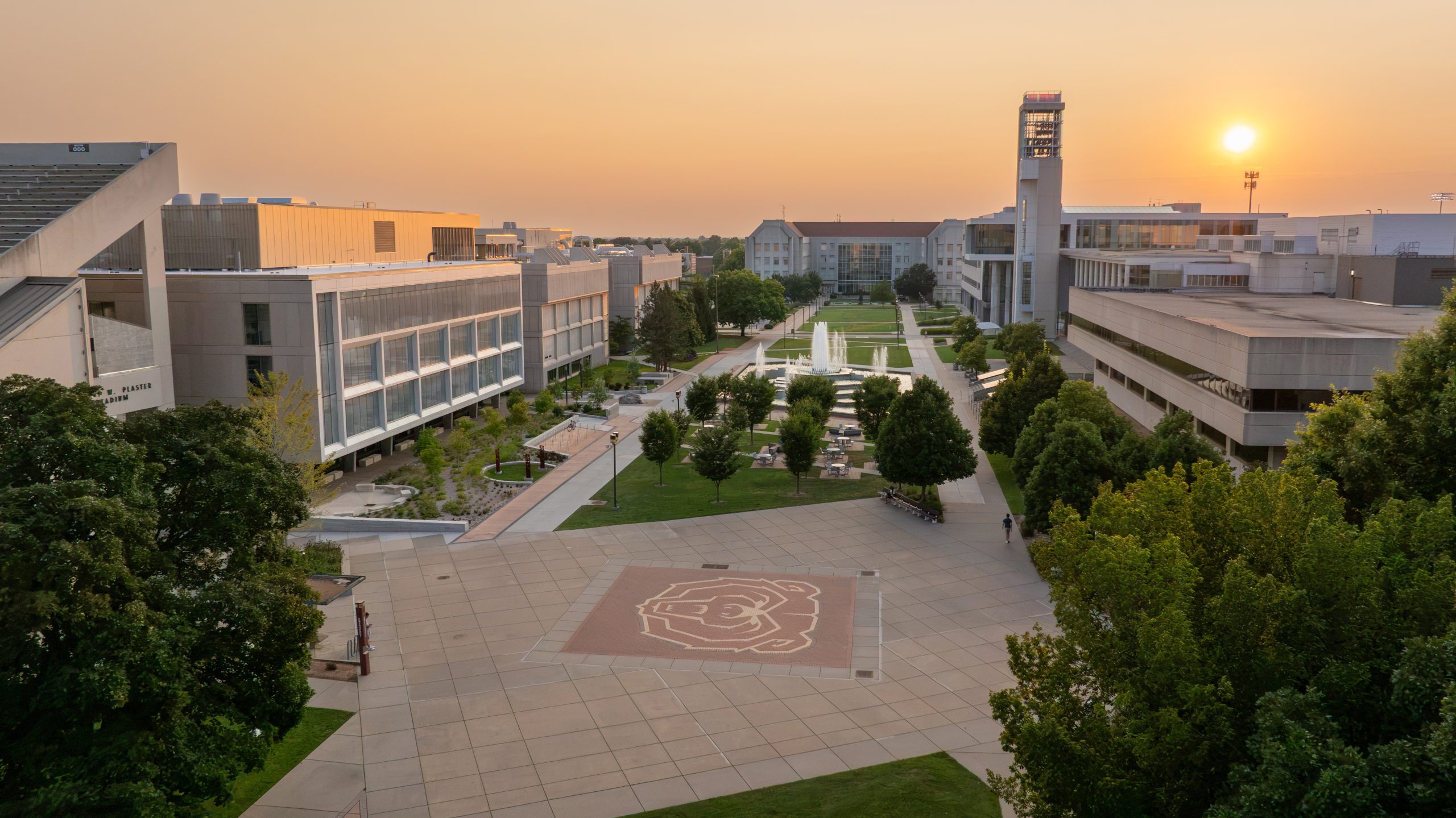 A campus plaza at sunset, featuring a large bear logo on the ground, surrounded by modern buildings and trees.