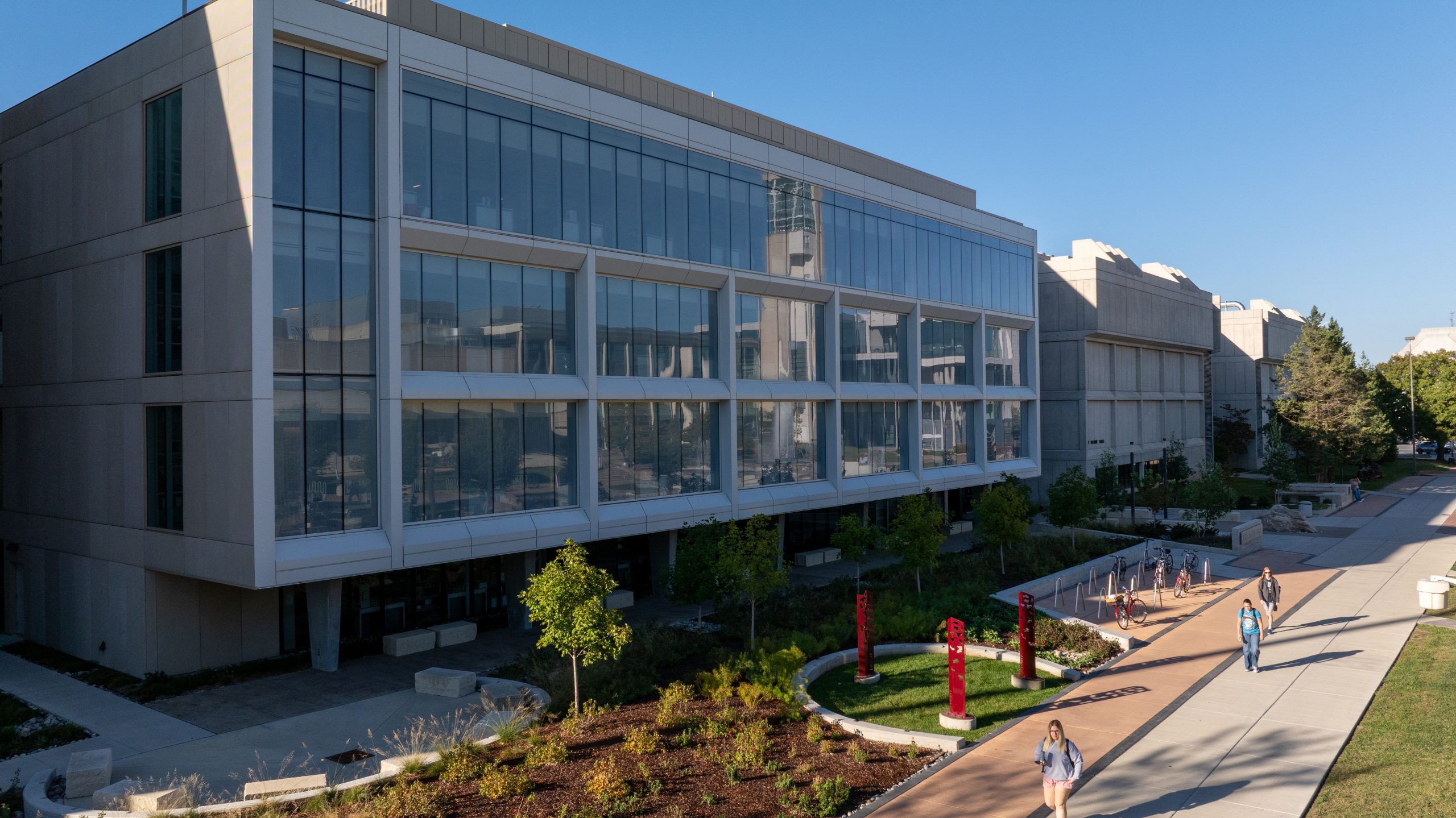 A modern university building with large glass windows and a landscaped walkway where students stroll under a clear blue sky.